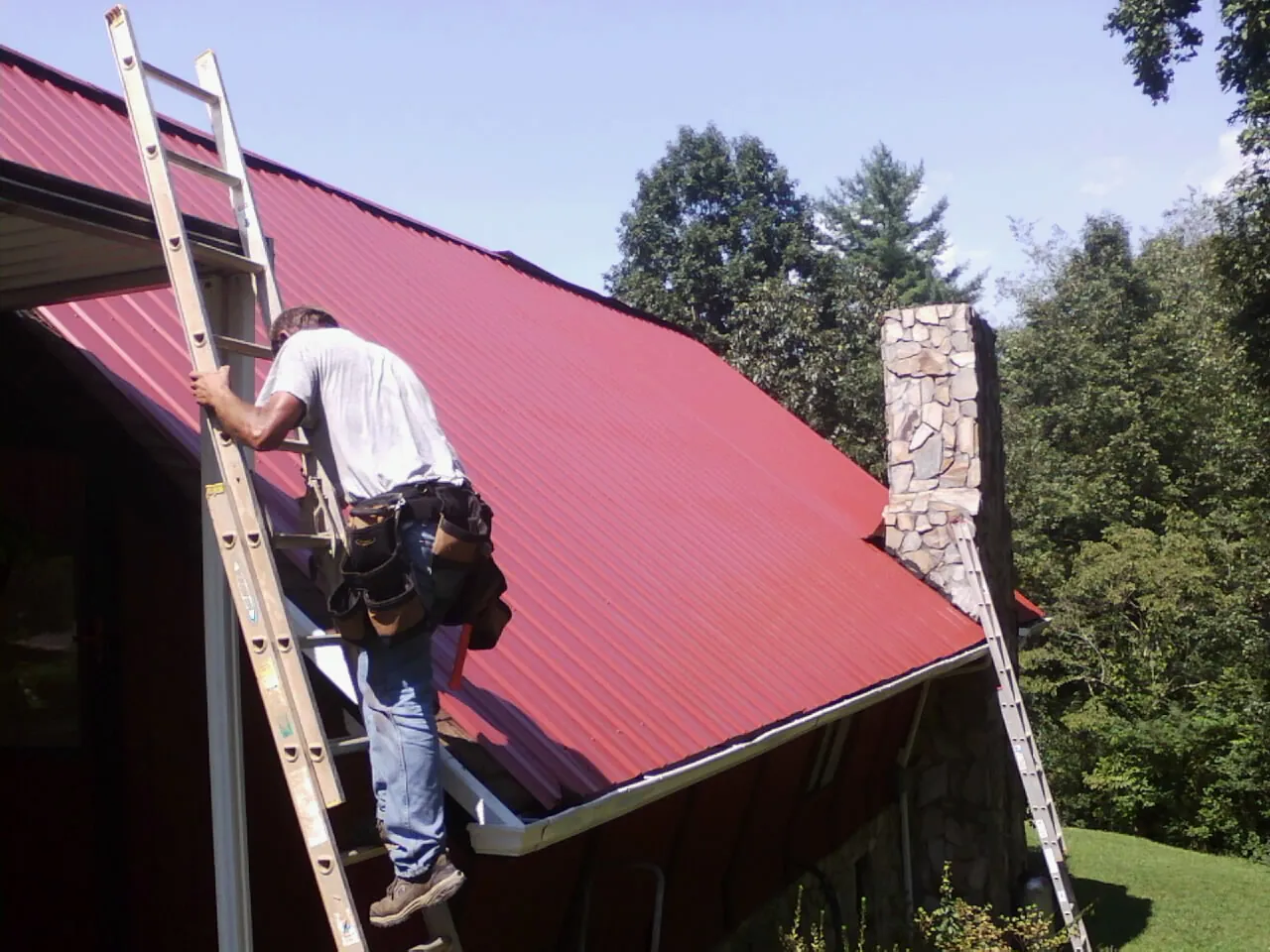 Roofer on a ladder beside a red metal roof and stone chimney — Soffit & Fascia Repair by Roofing Brothers in Frankford